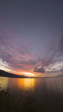 Arka planda dramatik bulutlar ve dağlarla gölün üzerinde gün batımı