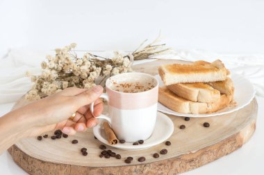 Kahve ve sütlü bardak, kapuçino, hafif arka planda tereyağlı kızarmış ekmekle servis ediliyor. Kahve fincanı alan kişi. Basit ve Portekiz kahvaltısı.