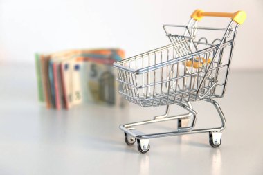 Boş alışveriş arabası ve para, beyaz arka planda birkaç avroluk banknot. Satın alma gücü, aile harcamaları, temel mallar, tüketici toplumu hakkındaki kavramsal imaj. Ön plan odaklı. Boşluğu kopyala