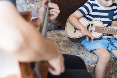 Beyaz çocuk küçük gitar veya ukulele ile müzik akorları çalıyor ve yapıyor, yakın çekim. Yetişkin biri çocuğa rahat bir şekilde öğretiyor..