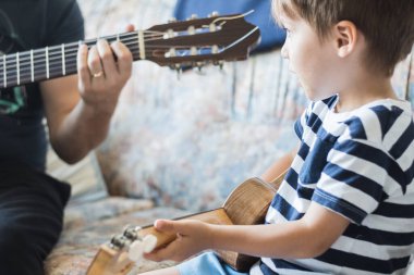 Beyaz çocuk küçük gitar veya ukulele ile müzik akorları çalıyor ve yapıyor, yakın çekim. Yetişkin biri çocuğa rahat bir şekilde öğretiyor..