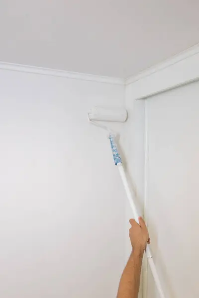 İşçi duvarları silindir ve beyaz boya kullanarak boyuyor, tadilat yapıyor, mekanları yeniden dekore ediyor. Dikey görüntü. Kapat ve boşluğu kopyala.