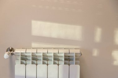 Sıcak güneş ışığı bir pencereden süzülerek beyaz bir radyatörün üstündeki bir duvara yumuşak gölgeler bırakıyor ve kapalı bir atmosfer yaratıyor..