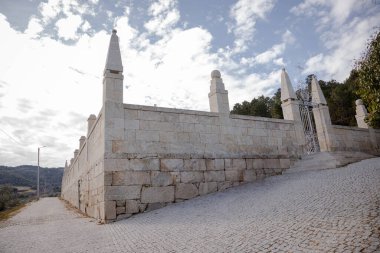 Tarihi bir mezarlığın taş duvarları ve sütunları. Parçalı bulutlu bir gökyüzünün altında demir bir kapı girişi var. Çakıl taşı yolu, heybetli geçide çıkıyor. Huzurlu ve zamansız bir atmosfer yaratıyor..