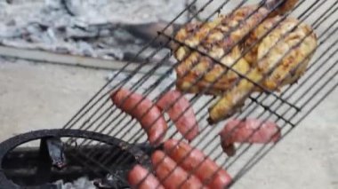 Yaşlı bir adam kamp ateşinin közleriyle sosis ve tavuk ızgara yapar, iştah açıcı ve geleneksel bir açık hava barbeküsü yaratır. Geleneksel BBQ kurulumu deneyimi.