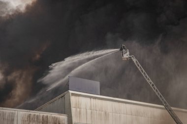 Bir itfaiyeci büyük bir sanayi yangınını söndürmek için hidrolik merdivenden su püskürtür. Yoğun siyah duman gökyüzüne yükselir ve durumun ciddiyetini ve tehlikesini gözler önüne serer.. 