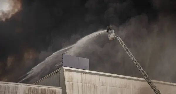 stock image A firefighter sprays water from a hydraulic ladder to extinguish a large industrial fire, with thick black smoke billowing into the sky, showcasing the intensity and danger of the situation. 