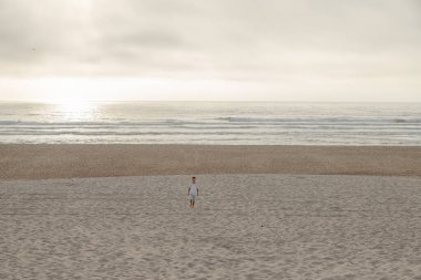 Çocuk gün batımında sakin bir plajın beyaz kumlarında yalınayak yürüyor. Doğayla temasta eğlenceli bir an.