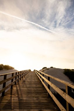 Güzel bir gün batımında, kum tepeleri arasında, yolu aydınlatan yumuşak altın bir ışıkla, tahta bir tahta kaldırım. Huzurlu ve mükemmel bir atmosfer Günün sonunda huzurlu bir yürüyüş.