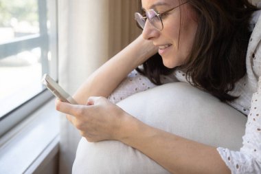 Gözlüklü bir kadın gün ışığında pencerenin yanında rahatlar, akıllı telefonuna bakarken gülümser. Evde sakin ve rahat bir an ve insanları yeni teknolojilere bağlamak.