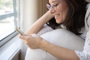 Gülümseyen kadın pencerenin kenarında rahatlıyor, akıllı telefonuyla kayarken neşeli bir anın tadını çıkarıyor. Neşeli ifadesi bağlılığı ve sıcaklığı yansıtıyor..