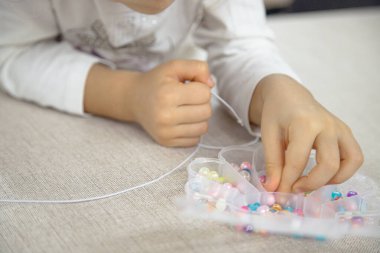 A young child carefully selects beads from a container to thread onto a string, demonstrating focus and creativity in making a colorful DIY jewelry piece. clipart