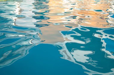 Gün batımında bir yüzme havuzunun yüzeyindeki soyut yansımalar. Dalgalanan desenler, gökyüzü, bulutlar ve ışığı sıvı bir bileşime harmanlayarak dingin ve büyüleyici bir görsel etki yaratır..