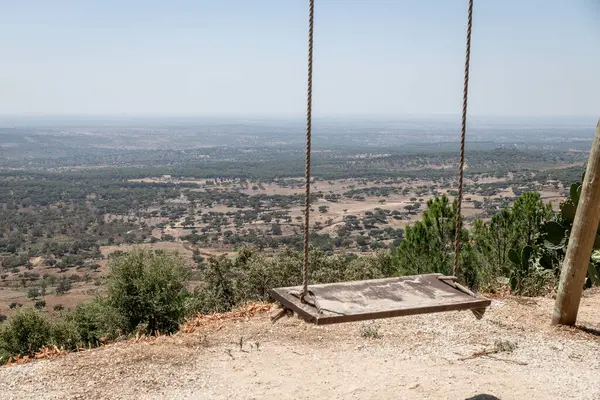 Güneşli bir yaz gününde, Carreiras, Portalegre, Alentejo, Portekiz 'deki Fonte dos Carvoeiros perspektifinde asılı boş ahşap salıncak, engin manzaranın huzurlu bir panoramik manzarasını sunuyor..