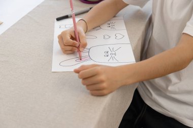 Bir çocuk, evde eğlenceli ve yaratıcı bir etkinlik sırasında tavşan, havuç ve kalplerin Paskalya temalı çizimlerini izler ve renklendirir. İnce motor becerilerini geliştiriyor..