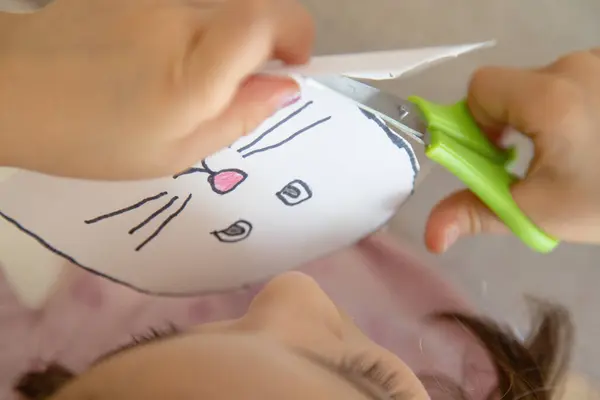 Evde, genç bir çocuk güvenlik makası kullanarak çizim hatlarını dikkatlice kesiyor, bir sanat etkinliği sırasında iyi motor beceriler, özerklik ve el kontrolü uyguluyor..