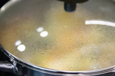 Ev yemeği yaparken cam kapaklı buhar tenceresinin yakın çekimi, su yoğunlaşması ve ısı yansımalarını gösteriyor, yemek hazırlarken modern bir mutfakta çekilmiş..