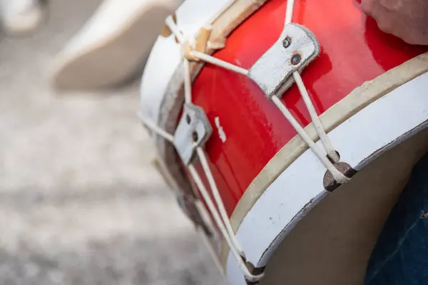 Açık hava kültürel etkinliği sırasında kırmızı ve beyaz ip gerilimli bas davul çalan bir kişinin yakın çekimi, geleneksel perküsyon detaylarını ve ritmik performans enerjisini yakalıyor.