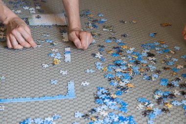 Gündüz vakti evdeki halı döşeli zeminde yapboz yapan bir kişinin elleri, odaklanmayı, sabrı ve kaliteli zamanı teşvik eden sakin bir aktiviteyi yansıtıyor..