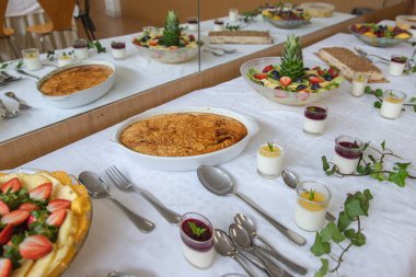 Çeşitli ev yapımı tatlılar ve taze meyveler, dekore edilmiş bir yemek alanında, muhtemelen bir kutlama ya da aile etkinliği olan kapalı bir etkinlik sırasında beyaz bir masa örtüsü üzerinde güzelce düzenlenir..