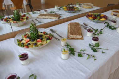 Çeşitli ev yapımı tatlılar ve taze meyveler, dekore edilmiş bir yemek alanında, muhtemelen bir kutlama ya da aile etkinliği olan kapalı bir etkinlik sırasında beyaz bir masa örtüsü üzerinde güzelce düzenlenir..