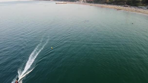 スローモーション英国ボーンマスビーチと海で高速移動スポーツボートの高角度空中映像 夏の美しい暑い日 人々は夏休みを楽しんでいる — ストック動画