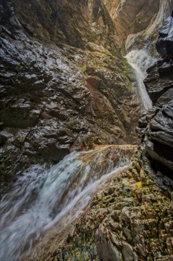Bir şelalenin güçlü akışı parlak ıslak taşlarla çevrili derin kayalık bir vadiyi keser.