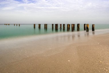 Napoli İskelesi ve sulara uzanan ahşap direklerle sakin bir deniz manzarası, parlak mavi gökyüzünün altında, zamansızlık ve sükunet hissi uyandırıyor..