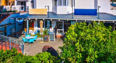 Mavi sandalyeli ve masalı açık bir kafe sahnesi, merkezi bir tezgah, bir menü tahtası ve yapraklı yeşillik. Rahatlamış bir titreşim yayıyor. Eğim kaydırma lensi minyatür bir görünüm yaratır