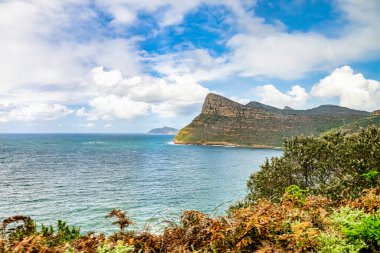 Heybetli kayalık uçurumlar, yemyeşil yapraklar ve bulutlu bir gökyüzünün altında sakin mavi deniz manzarası mükemmel bir manzara kaçışı sağlıyor..