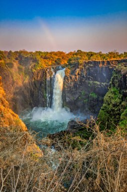 Çarpıcı bir şelale, canlı bir gökyüzünün altındaki havuza dökülür. Doğanın saf ve güçlü güzelliğini canlı renklerde gösterir..