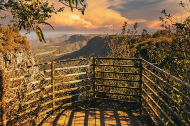 A captivating view of a sunlit mountain landscape from a rustic wooden viewpoint, surrounded by lush greenery under a golden sky. clipart