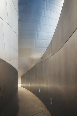 Çarpık metal paneller ve dramatik ışıklandırma altında dar bir geçitin yer aldığı modern bir mimari sahne, bir merak duygusunu uyandırıyor..