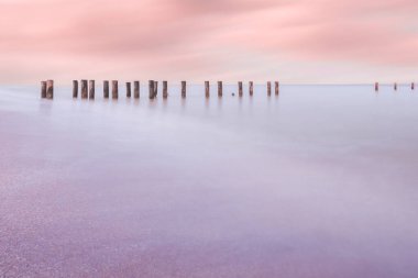 Durgun bir denize yansıyan pastel renkli gökyüzü ile rüya gibi bir deniz manzarası, yumuşak sularda hareketsiz duran ahşap direkler, huzuru ve yansımayı çağrıştırıyor..
