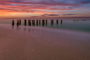 Nefes kesen bir günbatımı gökyüzünde canlı renkler bırakıyor ve sakin denizi yansıtıyor, tahtadan direkler önplanda sakin bir şekilde duruyor, Napoli Florida 'da huzurlu bir manzara yaratıyor.