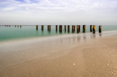 Napoli İskelesi ve sulara uzanan ahşap direklerle sakin bir deniz manzarası, parlak mavi gökyüzünün altında, zamansızlık ve sükunet hissi uyandırıyor..