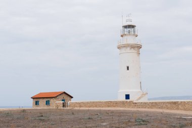 Beyaz deniz feneri ve arka plan olarak bulutlu gökyüzünün altında turuncu çatılı küçük bir ev.