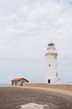 Beyaz deniz feneri ve arka plan olarak bulutlu gökyüzünün altında turuncu çatılı küçük bir ev.