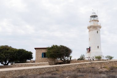 Beyaz deniz feneri ve arka plan olarak bulutlu gökyüzünün altında turuncu çatılı küçük bir ev.
