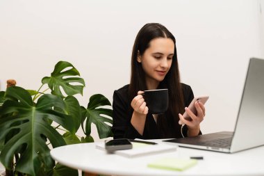 Ofiste bilgisayar başında çalışan beyaz duvarlı ve yeşil bitkili bir kadın kahve içiyor.