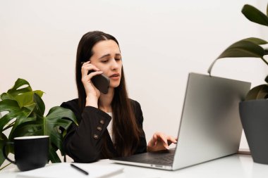 Ofiste bilgisayar başında çalışan beyaz duvarlı ve yeşil bitkili bir kadın kahve içiyor.