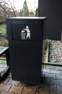 Siyah bir çöp tenekesi kaldırımın kenarında ahşap bir binanın yanında duruyor. Tasarımı bu açık ortamda uygun atık imhasını teşvik eder..