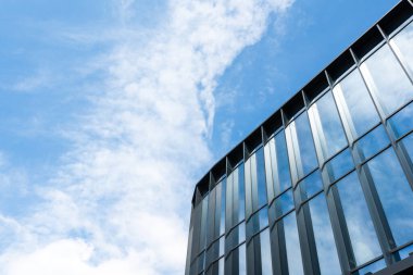 Parlak mavi gökyüzüne ve dağınık bulutlara karşı yansıtıcı pencereleri olan çağdaş cam bina cephesi. Mimari, iş veya kentsel gelişim kavramlarını resmetmeye elverişli.