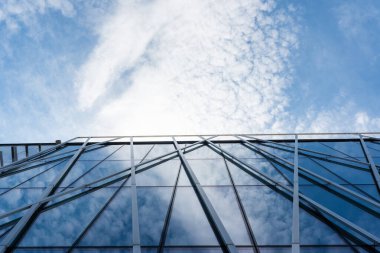Metal çerçeveli çağdaş cam cephe parlak mavi gökyüzünü ve dağınık bulutları yansıtır. Mimari, kentsel tasarım veya modern inşaat kavramları için uygun.