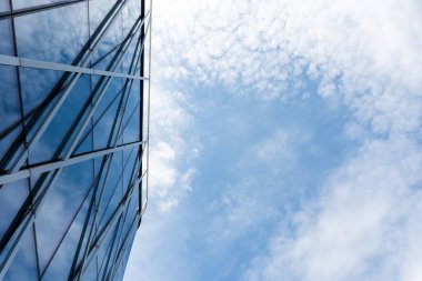 Yansıtıcı pencereleri ve geometrik çizgileri olan çağdaş cam bina cephesi dağınık bulutlu parlak mavi gökyüzüne karşı kurulmuş. Mimari, kentsel gelişim veya sunum ve pazarlamada iş kavramlarını resmetmeye uygundur