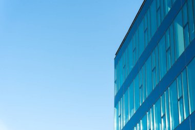 Çağdaş ofis binası, yansıtıcı cam pencereleri ve açık mavi gökyüzüne karşı kurulmuş temiz çizgileri var. İş, mimari veya kentsel gelişim kavramlarını resmetmeye uygundur.