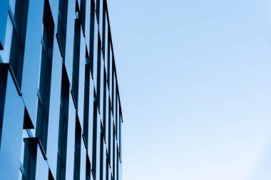 Yansıtıcı pencereleri ve temiz çizgileri olan çağdaş cam ofis binası, açık mavi gökyüzüne karşı kurulmuş. Kentsel mimari, iş ortamları veya modern şehir kavramlarını resmetmeye elverişli.