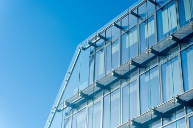 Çağdaş ofis binası parlak mavi gökyüzü altında yansıtıcı cam cepheli ve metal yapısal elementlere sahip. Mimari, iş, inşaat veya kentsel gelişim kavramlarını resmetmeye elverişli.