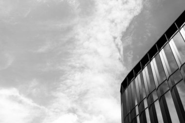 Siyah ve beyaz fotoğraf, yansıtıcı pencereleri ve dramatik bulutlu gökyüzü olan modern bir cam bina cephesini gösteriyor. Mimari, kentsel tasarım veya çağdaş iş kavramlarını resmetmeye uygundur.