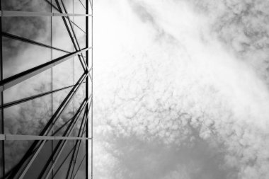 Modern cam bir binanın siyah beyaz fotoğrafı. Geometrik çizgiler bulutları yansıtıyor. Dramatik bir gökyüzüne karşı konumlandırılmış. Yaratıcı projelerde mimari, şehir tasarımı veya soyut arkaplan çizimi için uygundur.